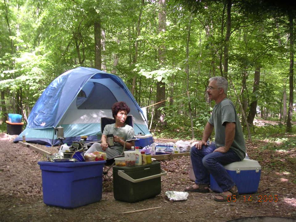Two people sitting around a campsite with a tent, coolers, and food
