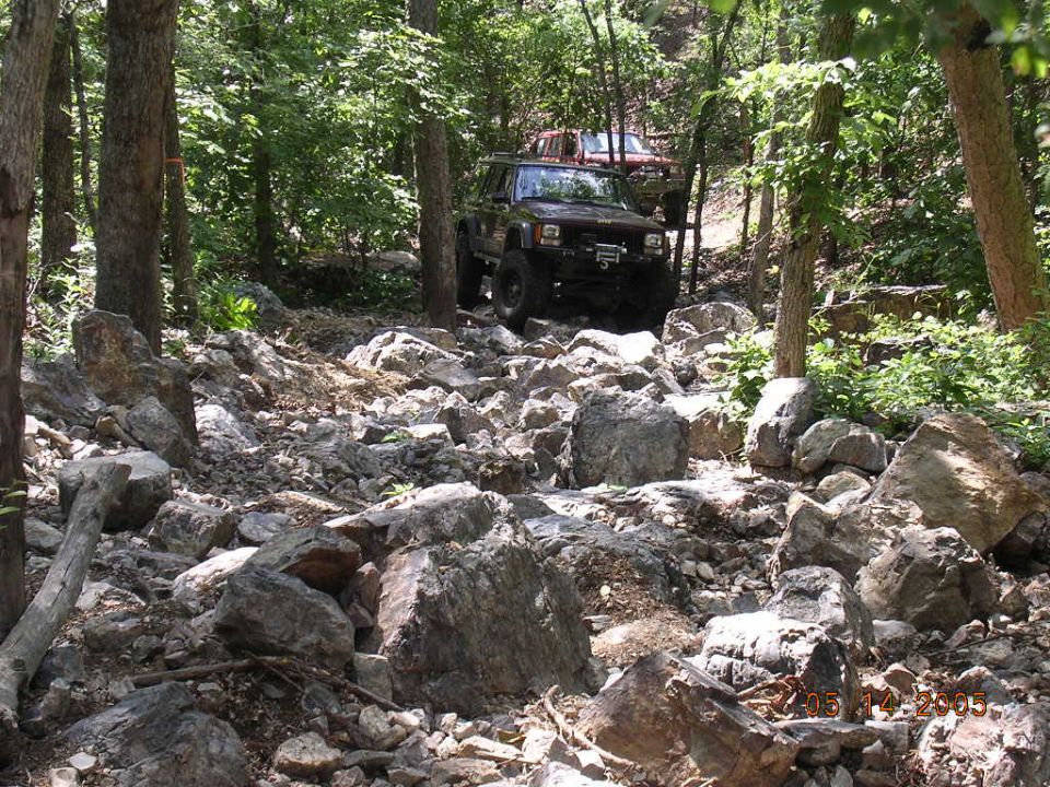Jeep Cherokee driving over rocks on a wooded trail. Another Jeep is behind it
