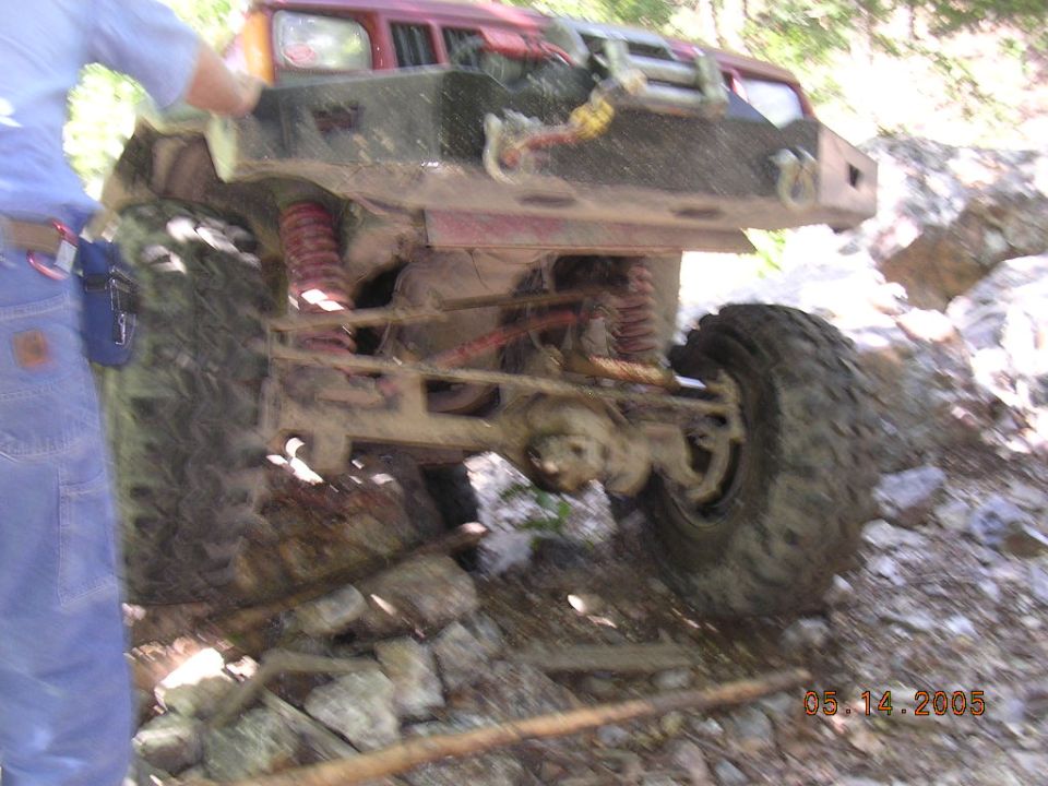 Red Jeep Cherokee with aftermarket bumper and winch, tires are large, on rocky terrain. Someone in blue jeans stands to the left