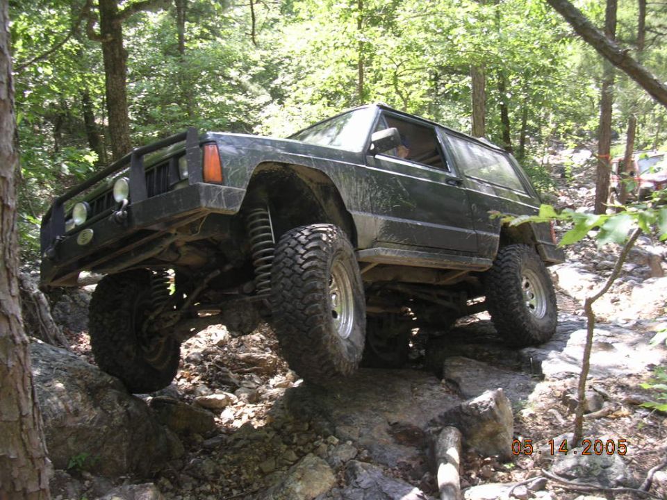 Black Jeep Cherokee climbing over rocks on a wooded trail. Another vehicle is visible in the background
