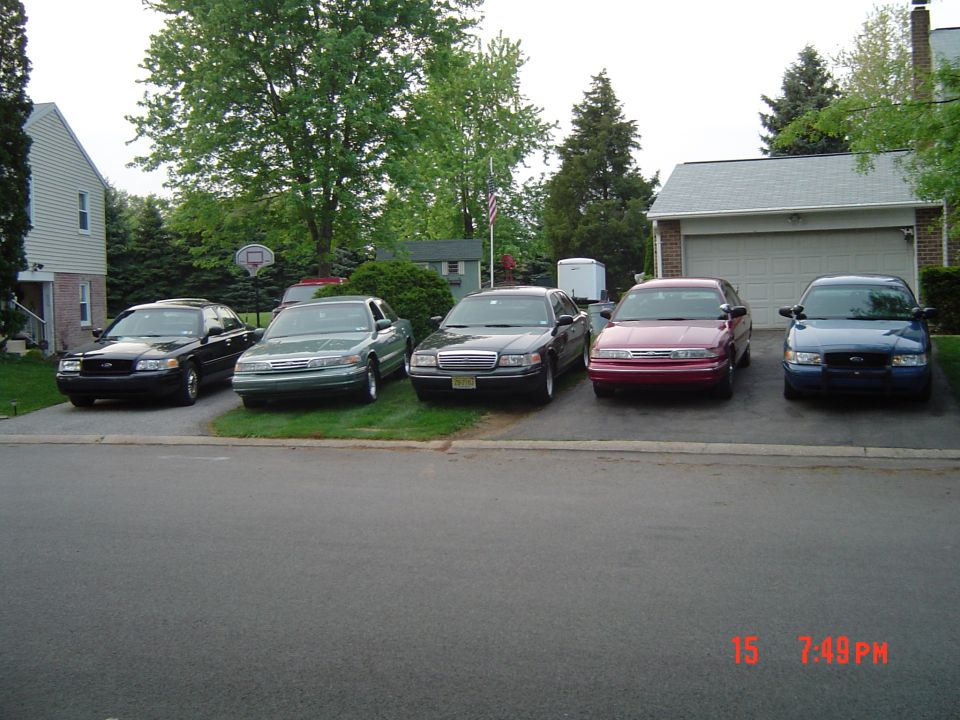 Five Ford Crown Victoria sedans parked in a driveway, varying colors including black, green, maroon, and blue. The front of each vehicle is visible