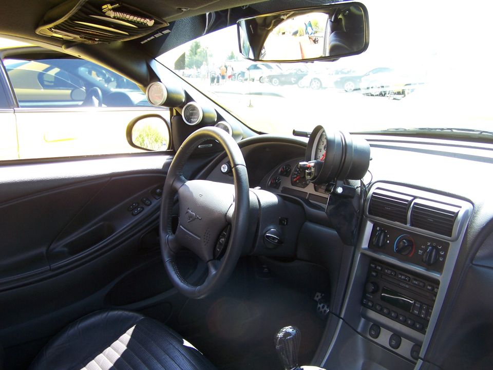Interior shot of a car with aftermarket gauges on the dash and a Mustang logo on the steering wheel. Another car is visible through the window