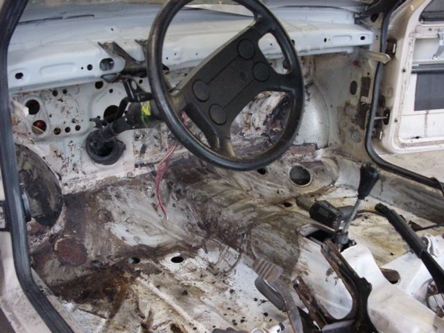 Car interior stripped down, showing bare metal floor, steering wheel, and gear shift. Appears to be from the late 80s or early 90s