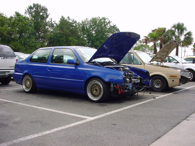 Blue 1998 Volkswagen Jetta with hood open and front end disassembled, parked next to other vehicles