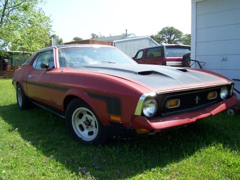 1971 Ford Mustang - A red 1971 Ford Mustang coupe with a black hood scoop and racing stripes is parked on grass next to a house, with another vehicle visible i…