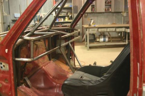 1990 Ford Bronco - stripped red truck cab fitted with a welded tubular roll cage and a black racing seat sits in a fabrication workshop.
