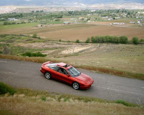 1996 Ford Probe - A red 1996 Ford Probe is captured from a high angle on a winding road with a scenic landscape in the background.