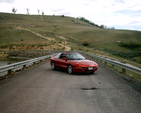 1996 Ford Probe - A red 1996 Ford Probe is captured in a three-quarter front view on a paved road, with a hilly landscape behind it.
