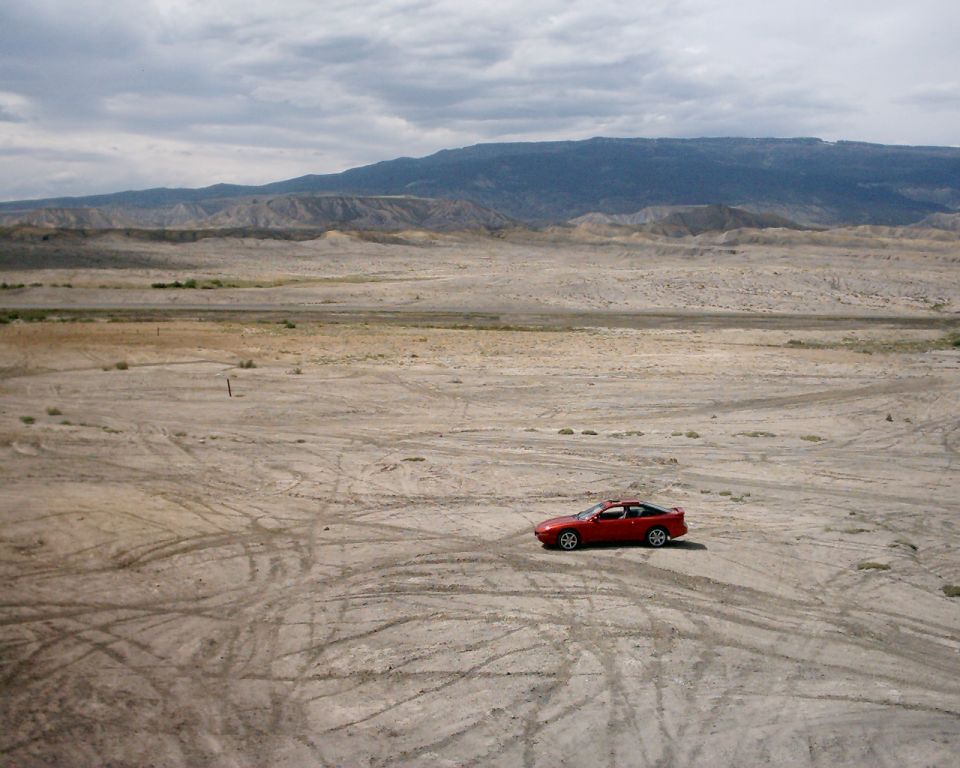 Red coupe parked alone on gray desert flats crossed with tire tracks below badlands and a mesa