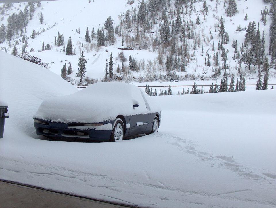 blue coupe buried in snow, snowy mountain and trees in background