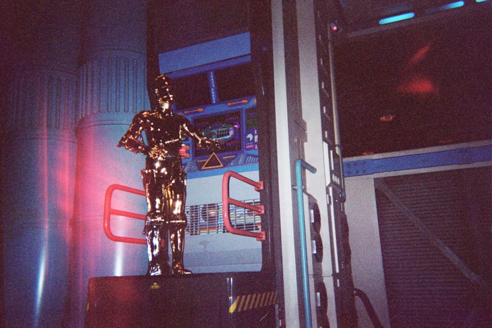 Gold C-3PO droid figure standing in a dimly red-lit themed ride queue or attraction set