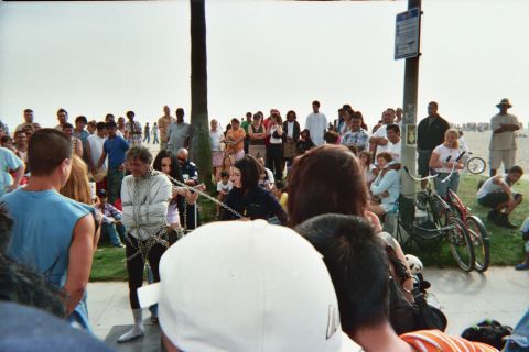 1968 Ford Mustang - A crowd watches a person in chains on a boardwalk, with palm trees and the ocean in the background.