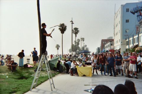 1968 Ford Mustang - A street performer juggles on a ladder at Venice Beach, with many people gathered to watch.