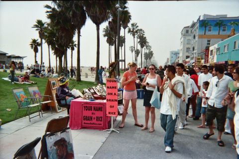1968 Ford Mustang - A lively scene unfolds on the Venice Beach boardwalk, with pedestrians strolling past shops and a vendor offering personalized rice grains.
