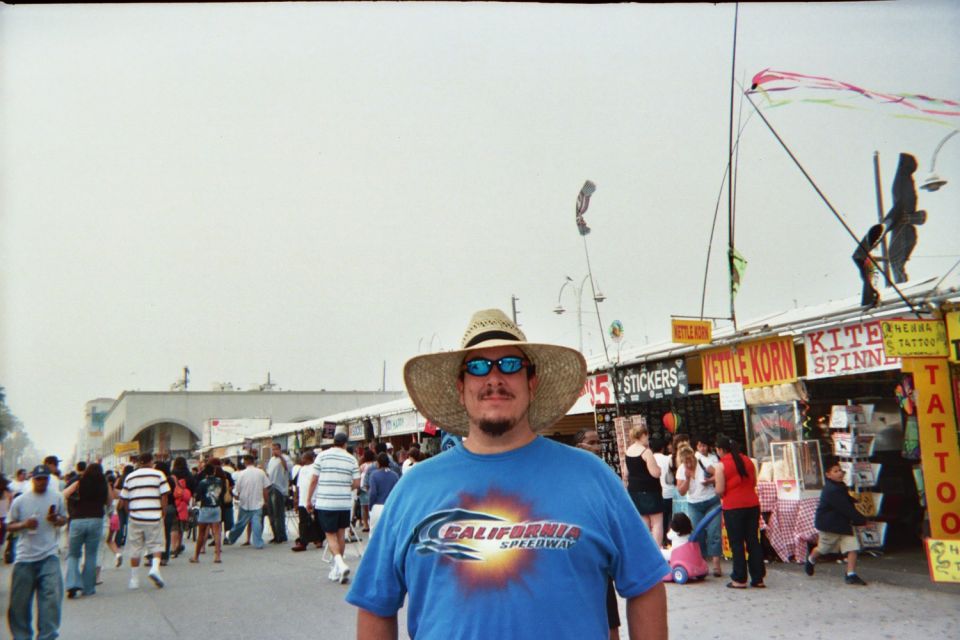 Man in straw hat and blue t-shirt with 'California Speedway' logo, standing in a busy outdoor market with stalls