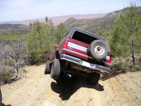 1989 Ford Bronco - Rear three-quarter view of a burgundy and silver two-tone Ford Bronco tipped up at a steep angle on a rutted dirt hill trail, driver-side r…