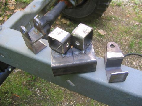 1978 Ford Bronco - Fabricated bare-steel suspension brackets laid out on a gray painted frame rail outdoors, a welded pair of boxed link mounts on a plate, pl…