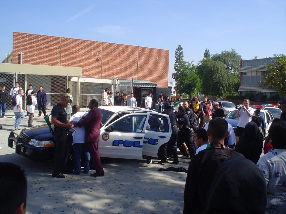 Black-and-white police cruiser with door open surrounded by officers and a crowd