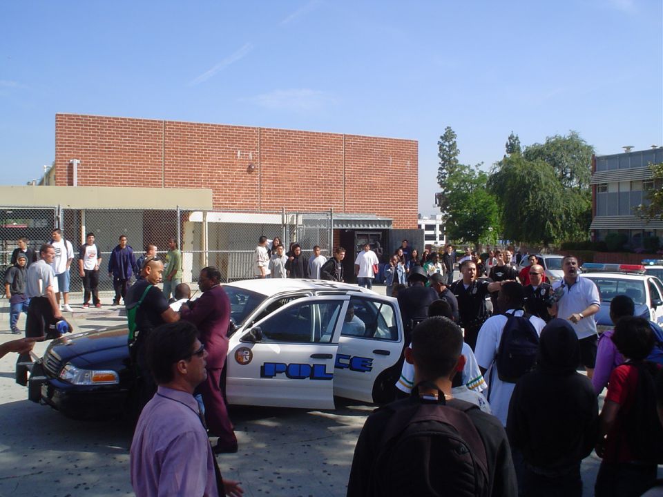 Police cars are parked outside a building with a crowd of people gathered around. One car has 'POLICE' written on the side