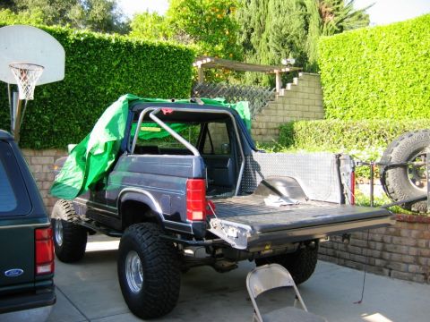 1985 Ford Bronco - This modified 1985 Ford Bronco features a roll cage and lifted suspension, partially covered by a green tarp.