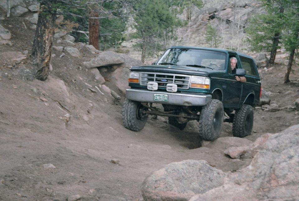 Green full-size 1990s Ford Bronco on big mud tires flexing over rocks on a forest trail