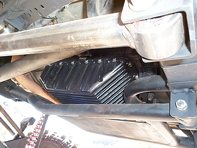 Black finned aluminum deep transmission pan seen from under a truck between frame rail and exhaust