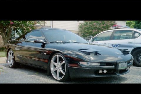 1995 Ford Probe - A black 1995 Ford Probe is parked next to a police car, its distinctive pop-up headlights and aftermarket wheels.