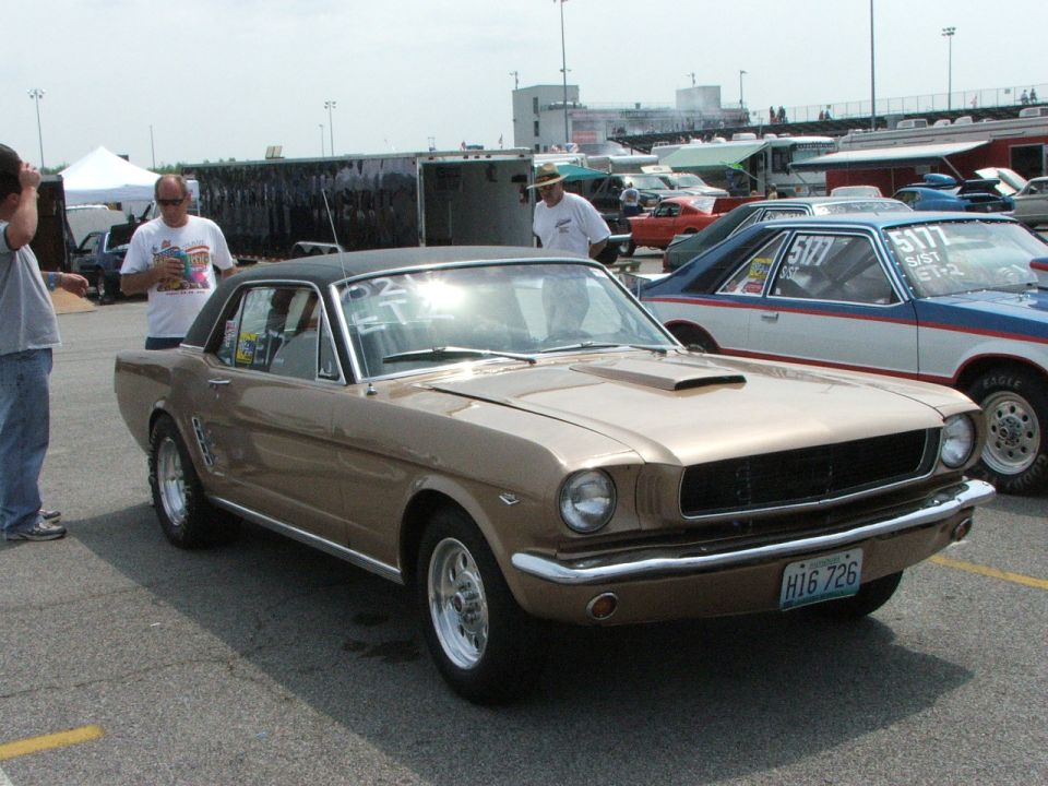 Gold classic Ford Mustang coupe with black vinyl top and hood scoop, 3/4 front view at a drag racing event