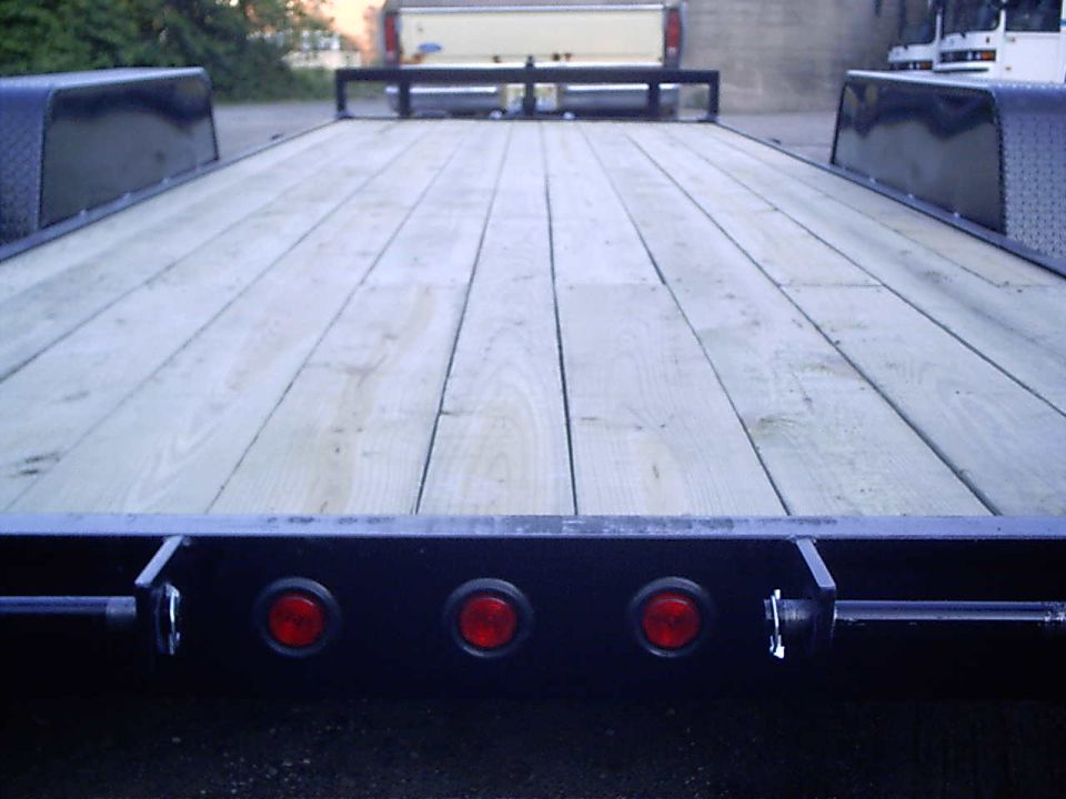 Flatbed trailer with wooden deck, three red lights on rear bumper. Pickup truck visible in background