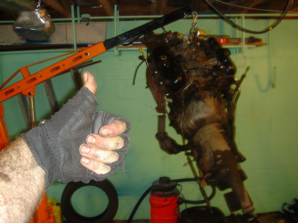 Engine and transmission assembly hanging from an engine hoist, with a gloved hand giving a thumbs up