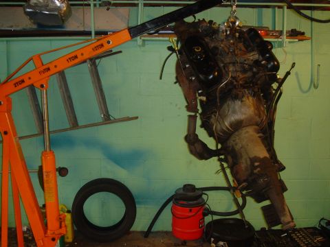 1986 Ford Mustang - An engine and transmission assembly is hoisted in a garage setting, with a tire and shop vac visible on the floor.