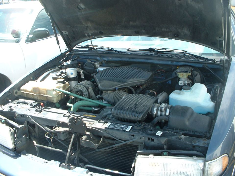 Engine bay of a dark blue car, hood open. Another white car is visible to the left