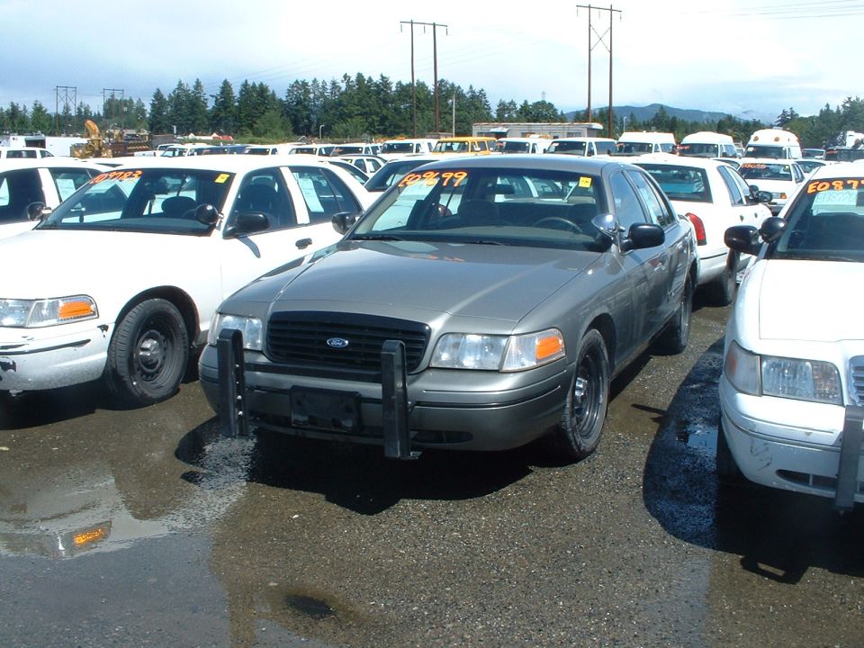 Gray 1999 Ford Crown Victoria police car with push bumper, parked in a lot with other white Crown Victorias