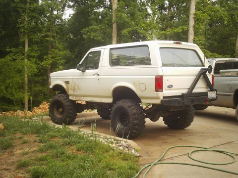 1996 Ford Bronco - A lifted 1996 Ford Bronco sits parked next to another vehicle, with a wooded area in the background.