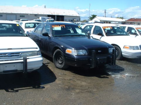 1999 Ford Crown Victoria - A dark blue Ford Crown Victoria police car is parked in a lot with several white vehicles under a bright sky.