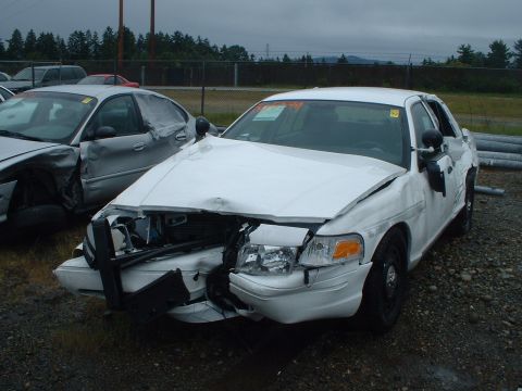 1999 Ford Crown Victoria - A damaged white sedan sits in a lot, its front end crushed, with another damaged car beside it.