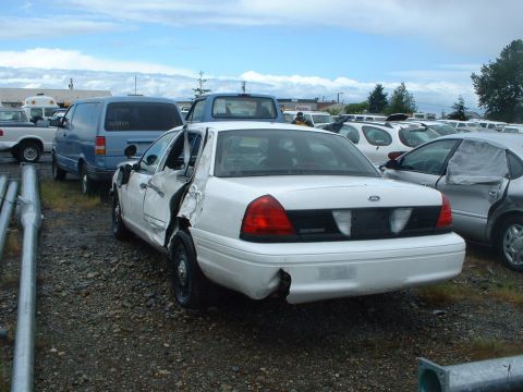 1999 Ford Crown Victoria - White sedan with significant rear quarter panel damage sits in a lot with other vehicles under a cloudy sky.