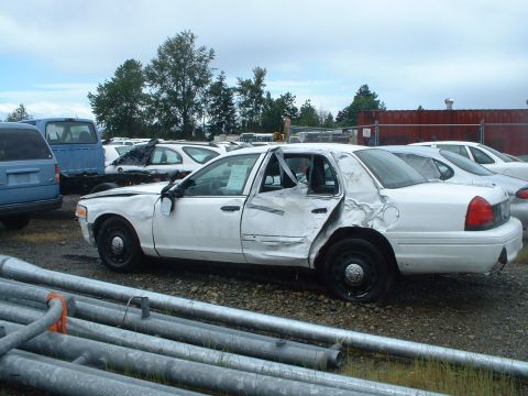 1999 Ford Crown Victoria - This damaged sedan is parked in a lot with other vehicles and some metal pipes in the foreground.