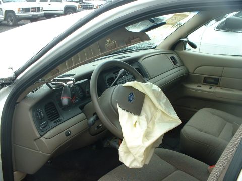1999 Ford Crown Victoria - The interior of a car shows a deployed airbag and the dashboard. Other vehicles are parked outside.