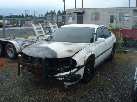 1999 Ford Crown Victoria - A damaged white sedan with a push bar sits in an outdoor lot next to a trailer.
