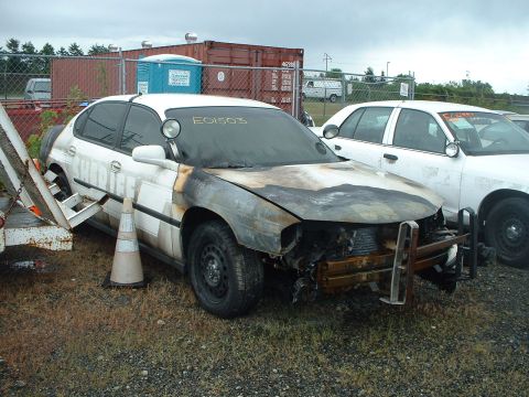 1999 Ford Crown Victoria - A severely damaged white sedan, possibly a former police vehicle, sits next to another white sedan in an outdoor lot.
