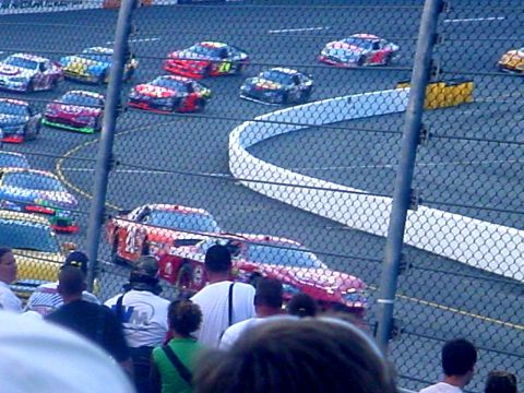 1973 Ford Ranchero - Multiple stock cars compete on a race track, seen from the spectator area behind a fence.