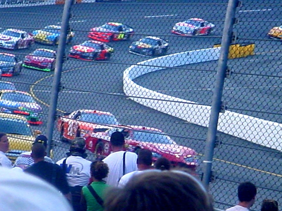 Stock cars racing on a track, viewed through a fence