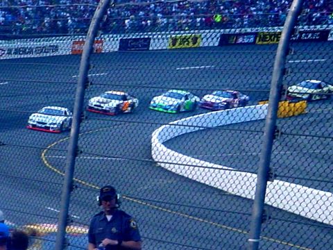 1973 Ford Ranchero - Multiple stock cars compete on a race track, viewed through a fence.