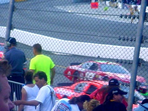 1973 Ford Ranchero - A red race car with visible racing decals and numbers is seen on the track from a 3/4 front perspective, with spectators in the foreground.