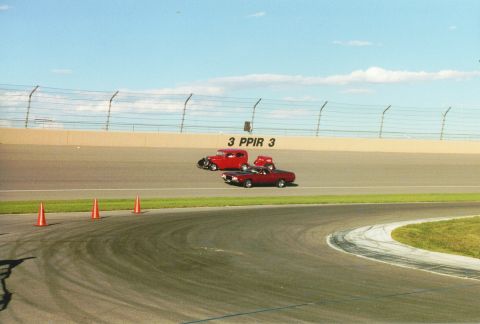1972 Ford Ranchero - Two red vehicles, a coupe and a pickup truck, compete on a racetrack under a blue sky with scattered clouds.