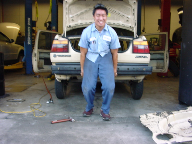 Man in blue shirt and pants lifting beige Volkswagen Jetta's rear bumper. The car's trunk is open