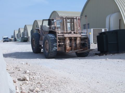 1988 Ford Bronco - A large forklift sits in a lot with several arched buildings and a truck in the distance under a clear sky.
