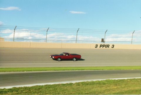 1972 Ford Ranchero - A red coupe utility vehicle with a black hood stripe drives on a race track under a blue sky.