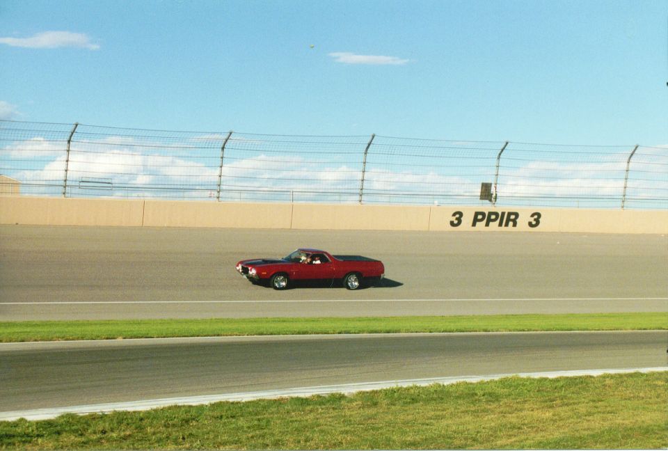 Red car-based pickup with black hood driving the banking of a speedway past a PPIR turn-3 wall